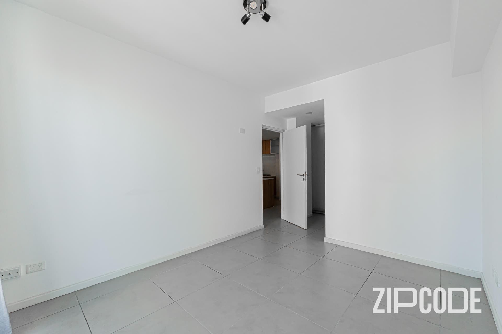 Dormitorio vacío con piso cerámico, puerta y pasillo de entrada.
