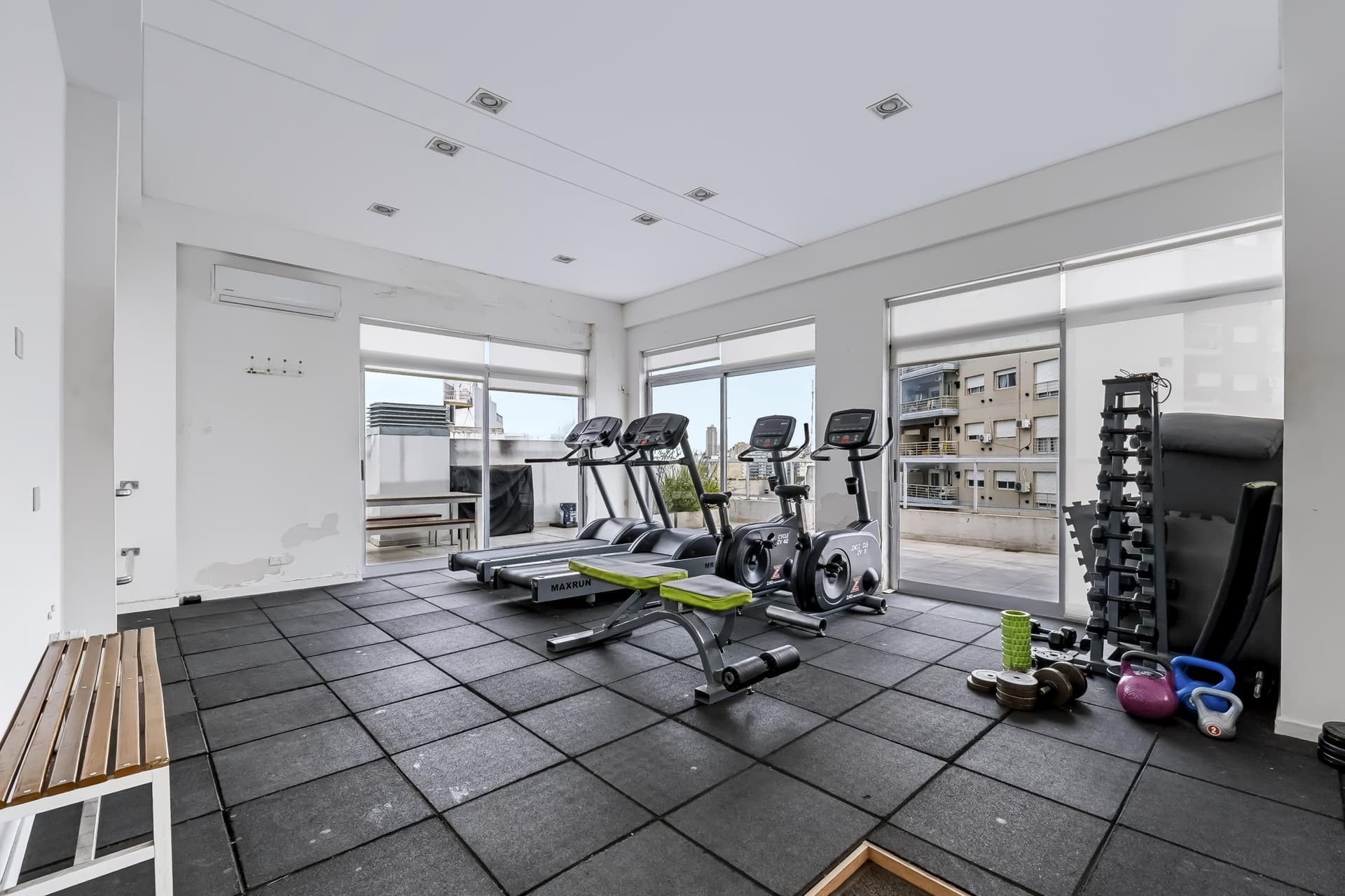 Gimnasio común con máquinas de ejercicio, vista exterior y luces empotradas.