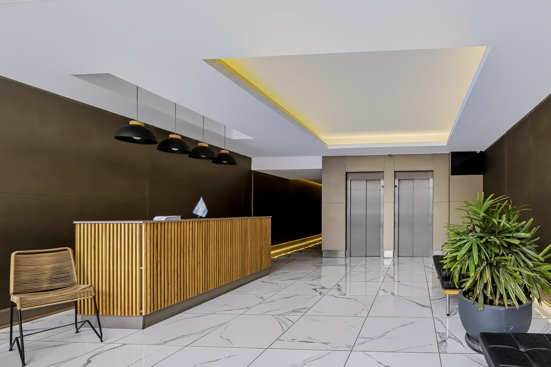 Recepción del edificio con mostrador de madera, ascensores y plantas decorativas.