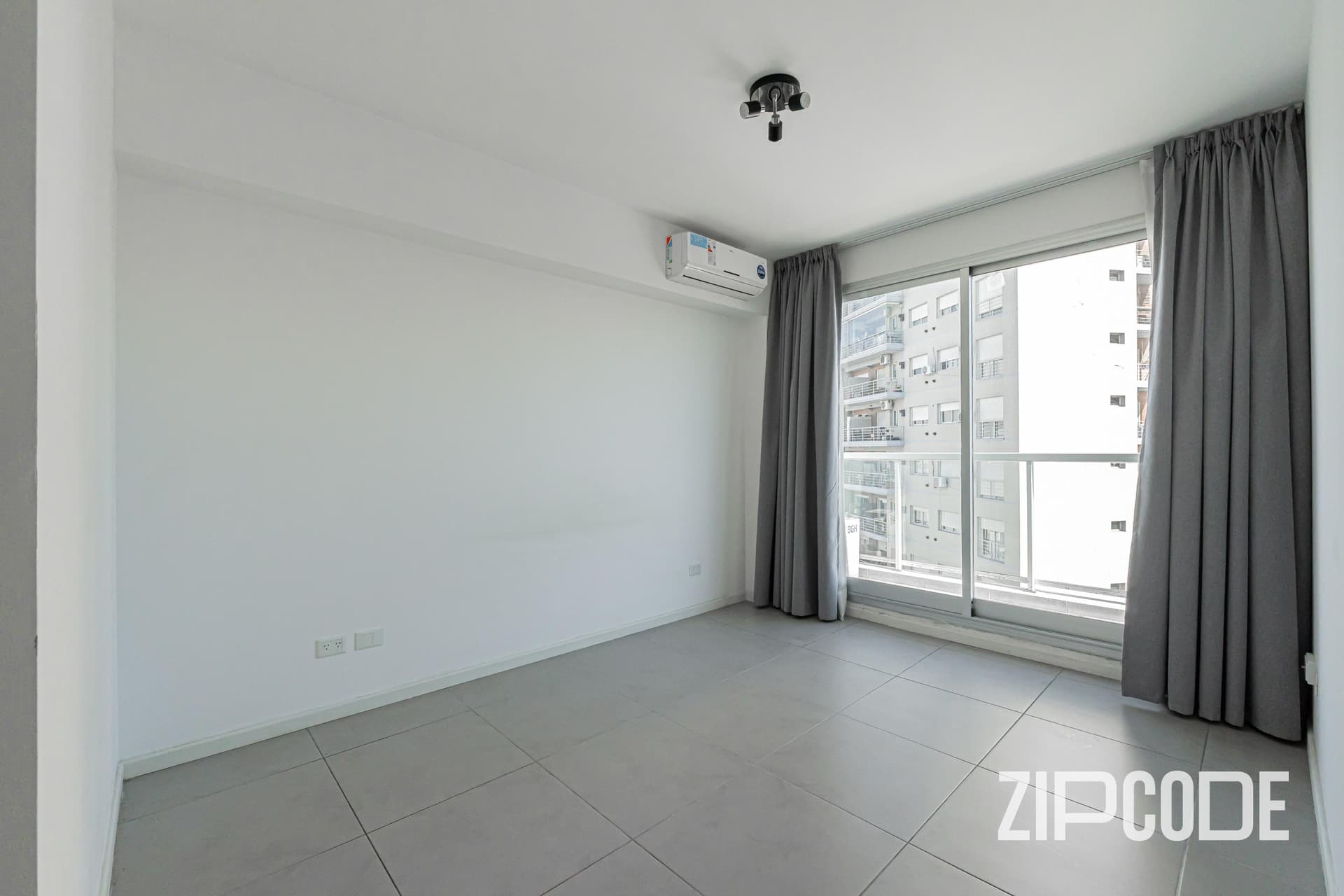 Dormitorio vacío con ventana, balcón y aire acondicionado en pared blanca.