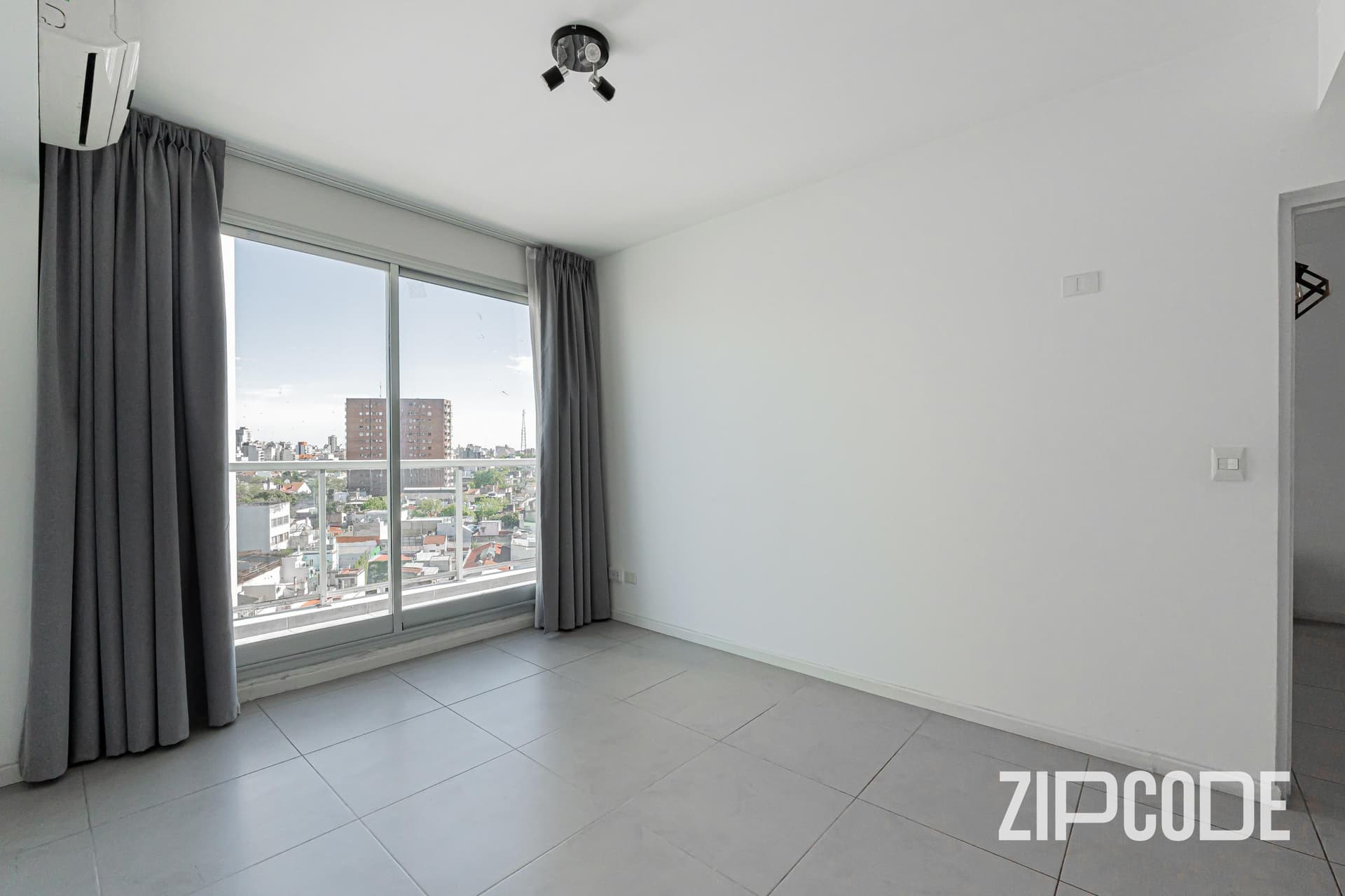 Dormitorio vacío con ventana grande, cortinas grises y piso cerámico.