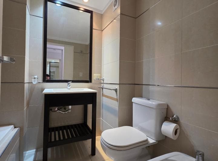 Baño con inodoro, bidet, vanitory con espejo y azulejos beige.