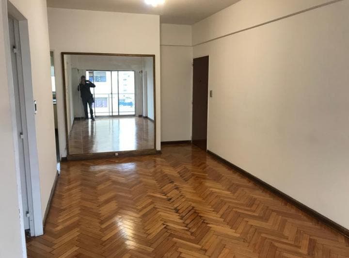 Living vacío con piso de parquet y espejo grande que refleja el ventanal.